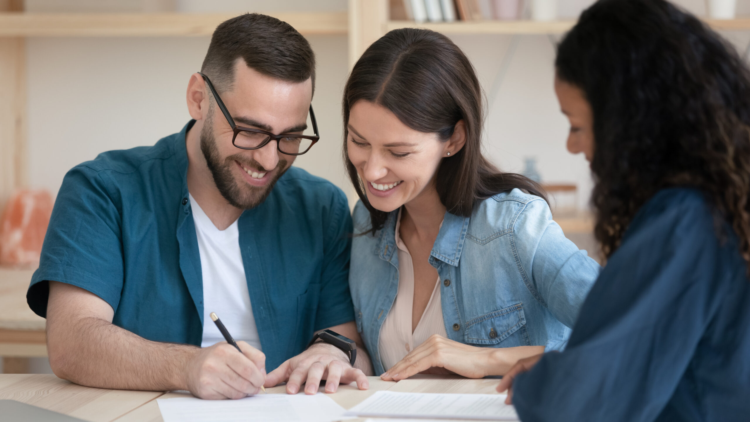 Two people seated together at a table signing a mortgage document while a loan officer presents a second page