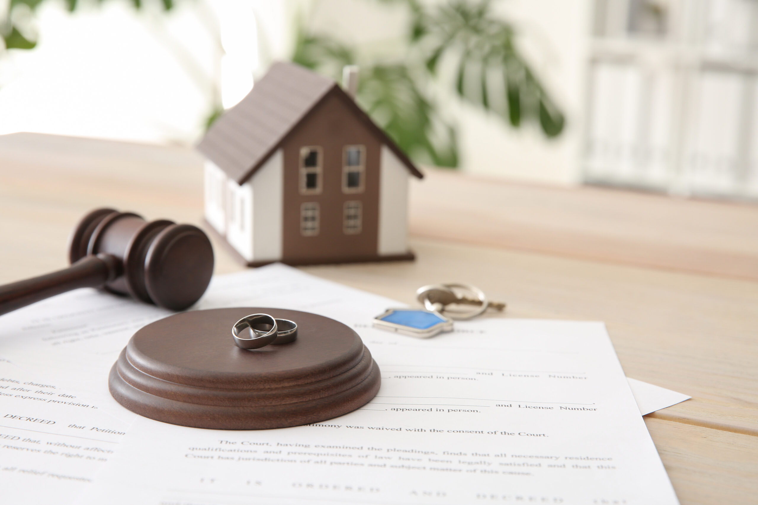 Wedding rings and a house key resting on a court decree beside a gavel and a model home on a wooden table