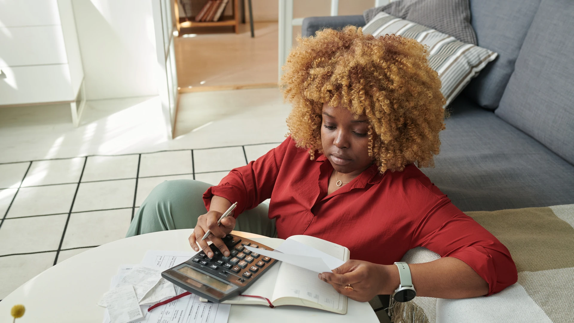 Woman adding up household bills on a calculator with receipts and a notebook spread on a coffee table