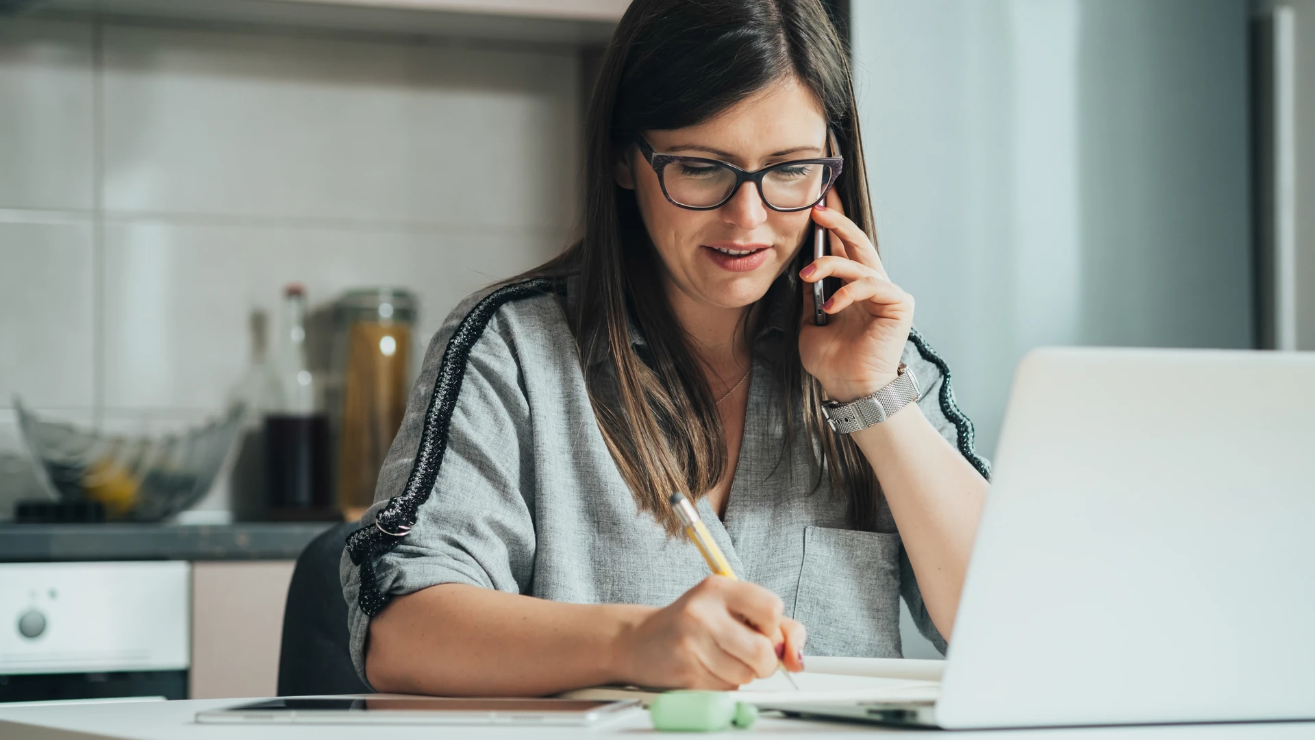 Woman on a phone call taking notes at her kitchen table beside an open laptop