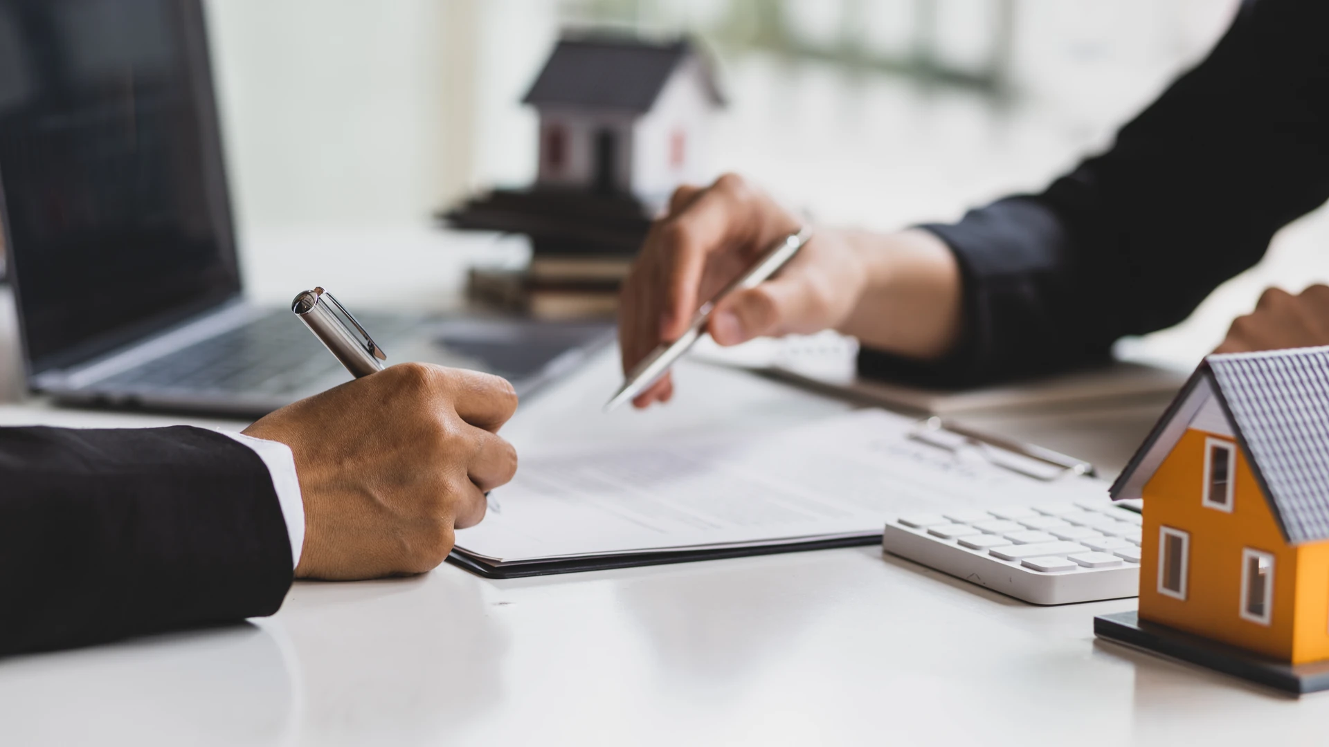Contact Us image by Fellowship Home Loans Two people's hands at a desk, one signing a document and the other pointing with a pen, beside a laptop, calculator, and small model houses