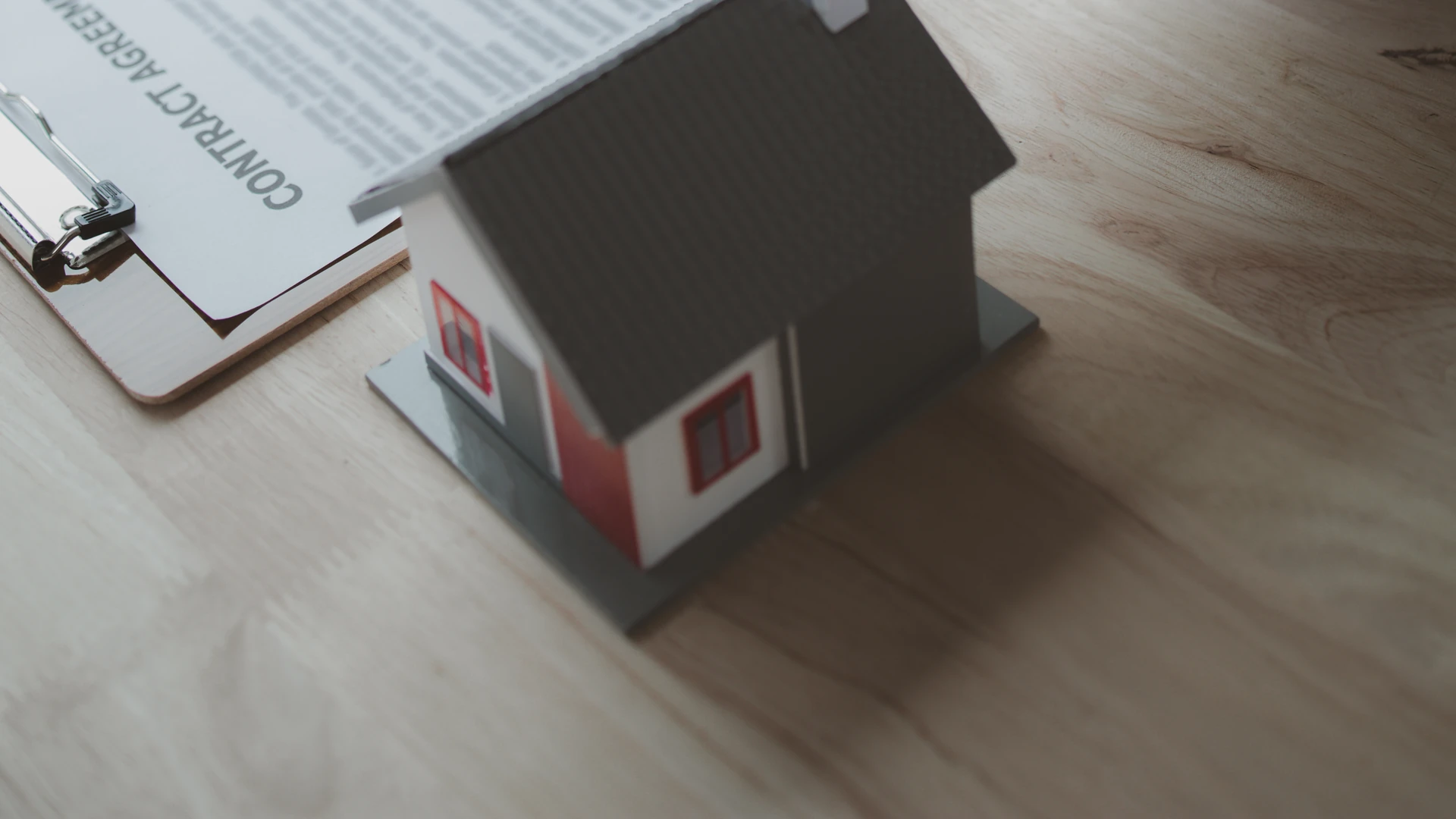 Contact Us image by Fellowship Home Loans A small model house beside a clipboard holding a document titled Contract Agreement, resting on a wood desk