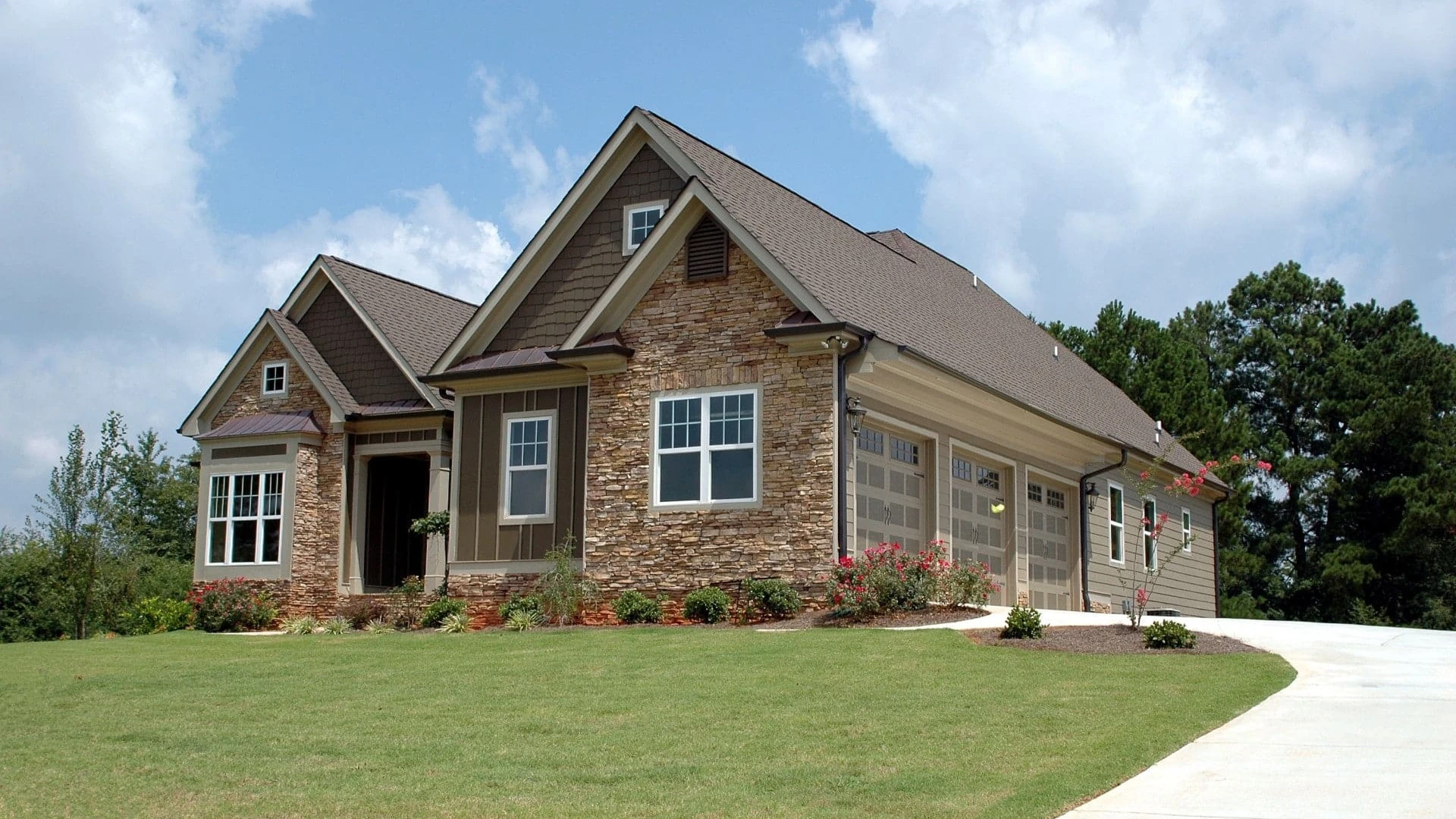 Contact Us image by Fellowship Home Loans Stone and siding house with gabled rooflines, a three-car garage, flower beds, a green lawn and a curved concrete driveway under a blue sky with clouds