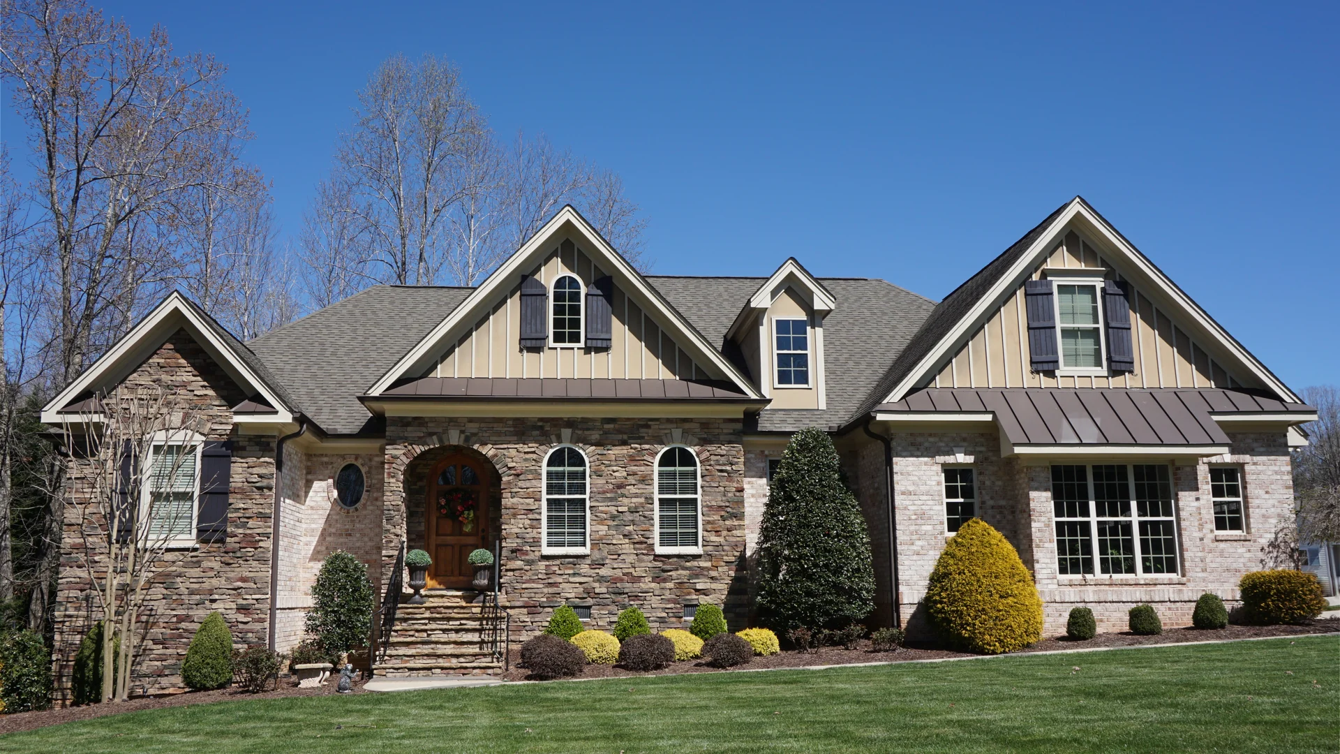 A house with a stone and brick front, board-and-batten gables with dark shutters, an arched wooden front door and trimmed shrubs behind a mown lawn under a clear blue sky