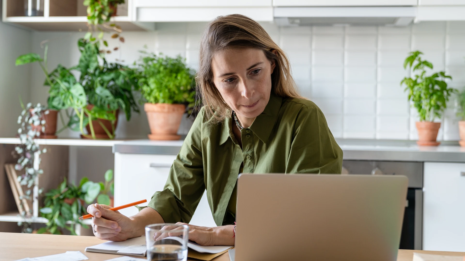 Woman in an olive green shirt holding an orange pencil over a notebook beside an open laptop at a table, with potted herbs and plants on shelves and a white tiled wall behind her