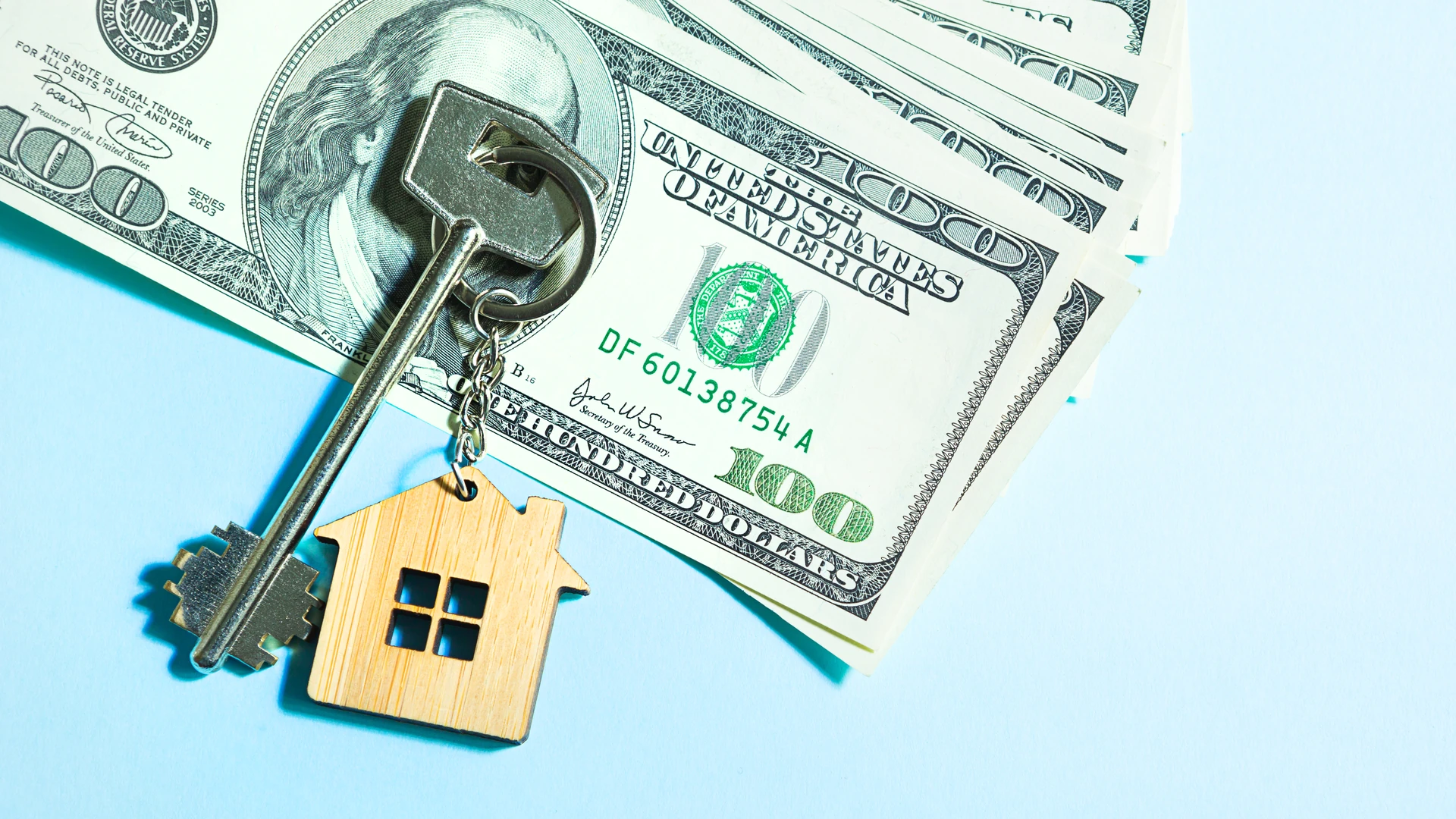 A silver key with a wooden house-shaped keychain lying on a fan of U.S. one-hundred-dollar bills against a light blue background