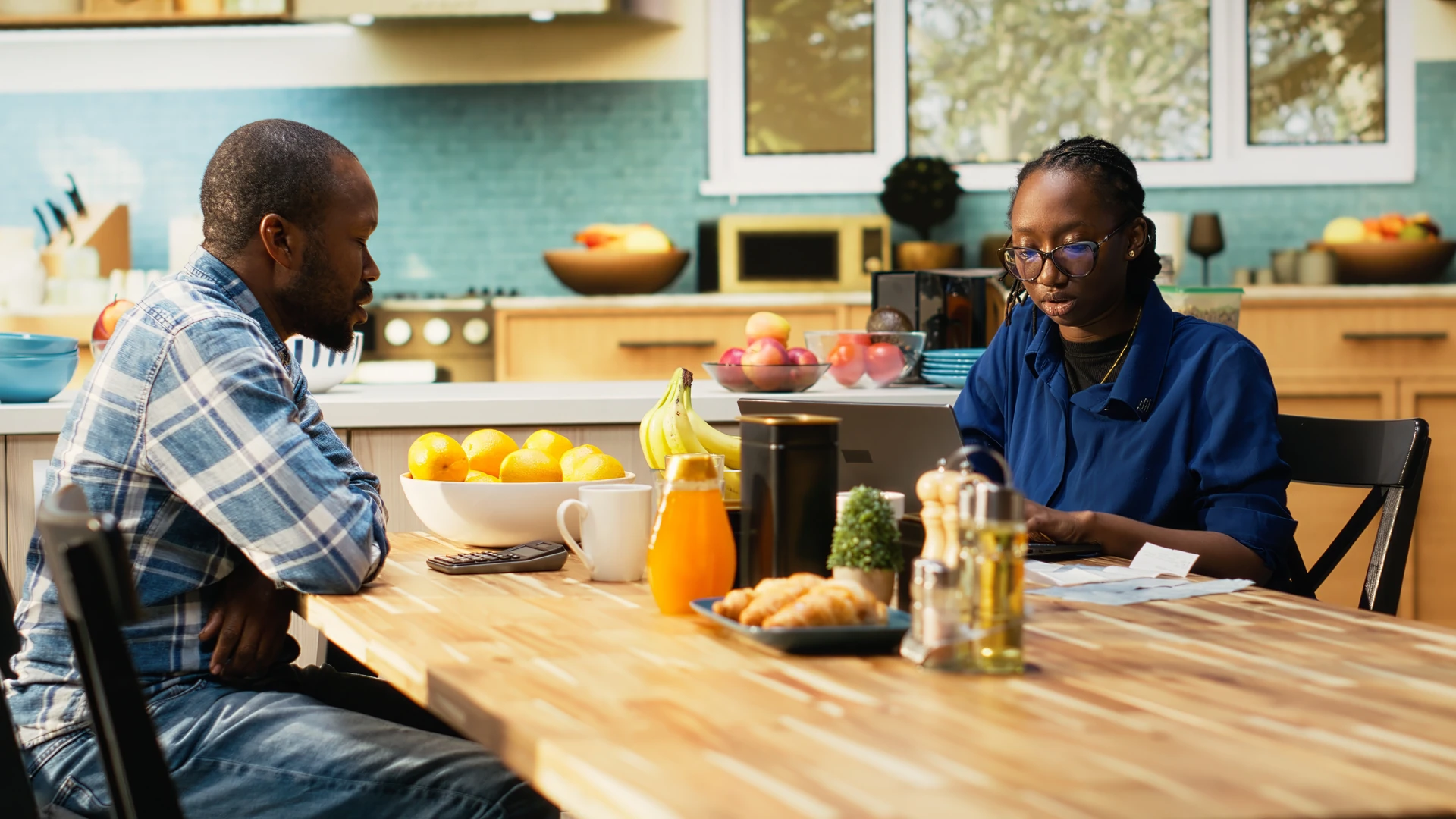 A man in a blue plaid shirt and a woman in glasses and a blue top sit at a wooden kitchen table with a calculator, receipts and a laptop, with a bowl of lemons and a plate of croissants nearby