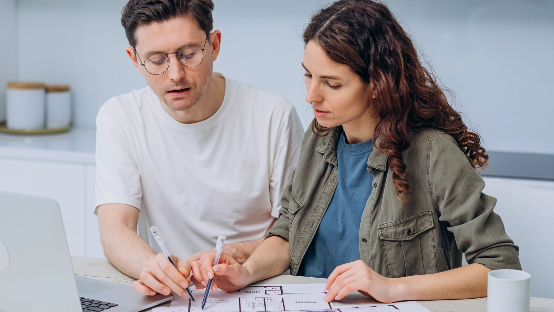 A man in a white T-shirt and glasses and a woman in an olive jacket hold pens over a printed floor plan on a table, with a laptop and a white mug beside them
