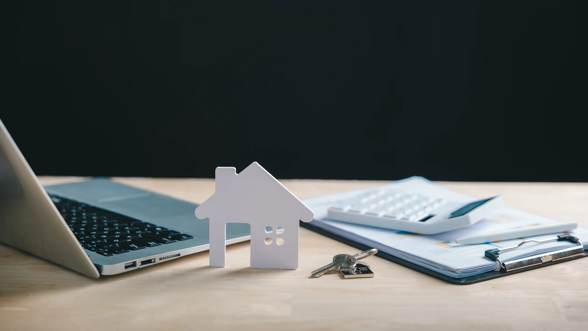 White paper house cutout and a set of keys on a wooden desk beside an open laptop, a calculator and a clipboard holding printed charts, against a black background
