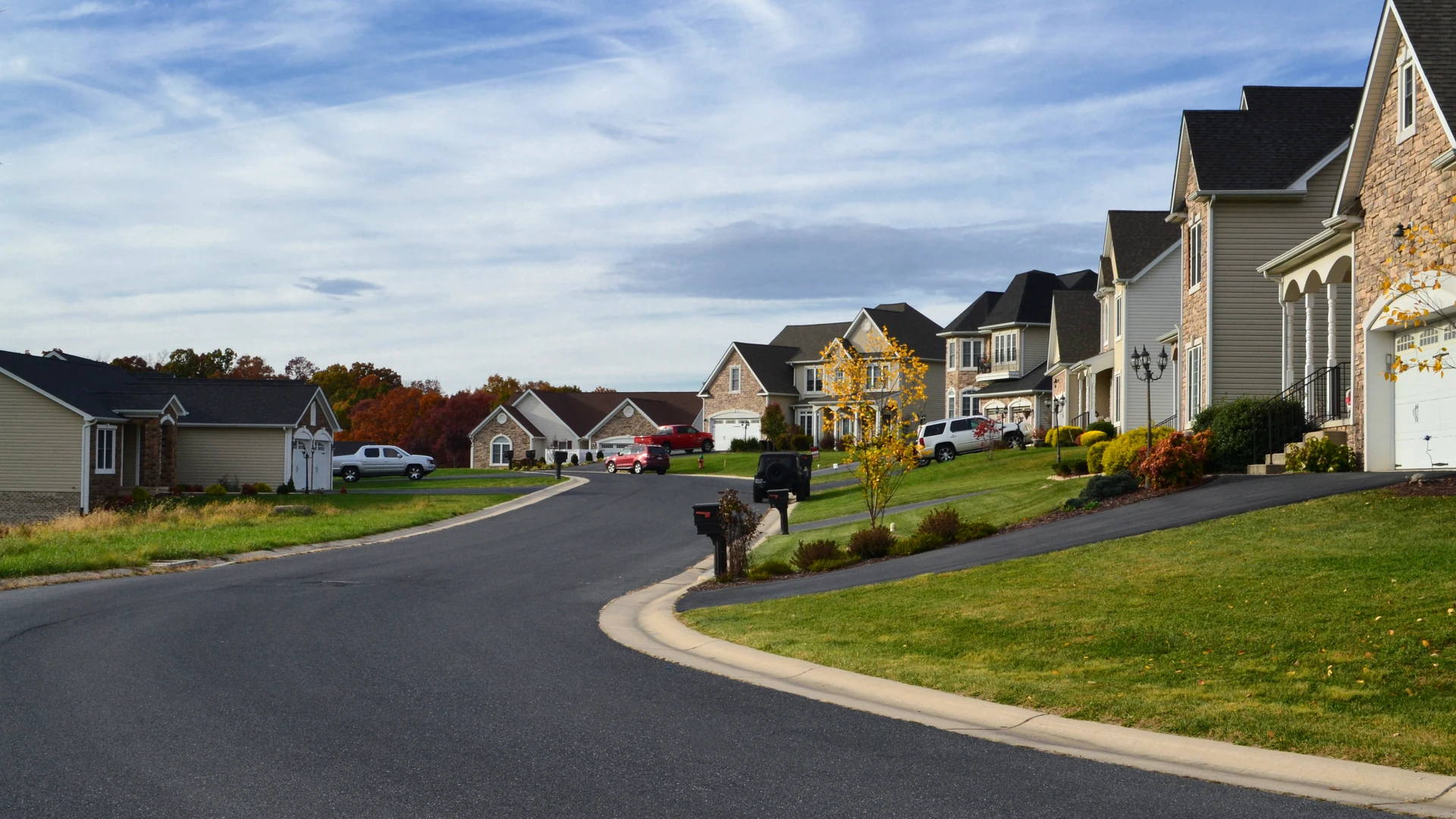Contact Us image by Fellowship Home Loans Curving suburban street lined with detached houses, driveways, mailboxes and green lawns under a partly cloudy sky, with autumn trees in the distance
