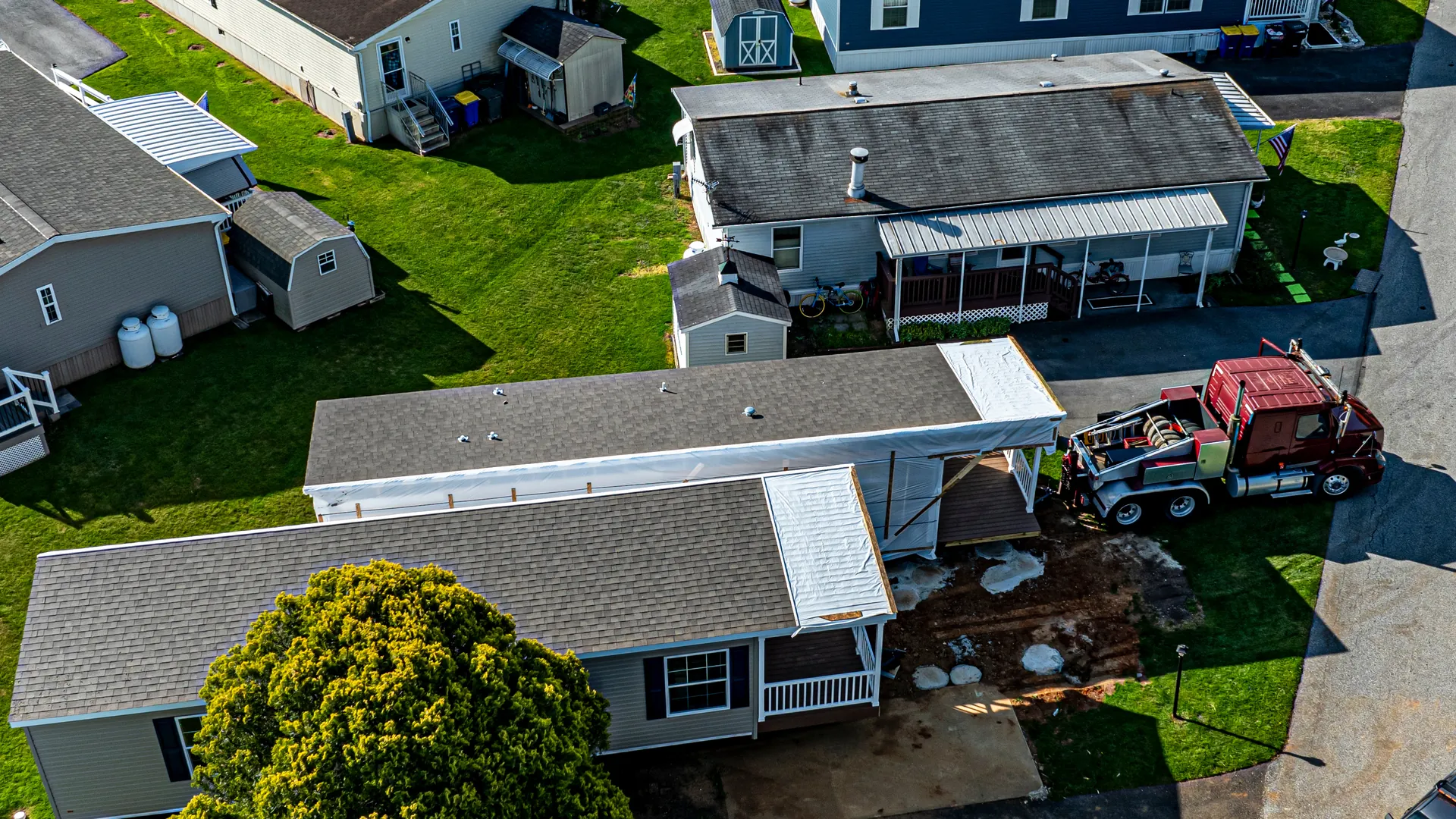 Aerial view of a manufactured home being set on its lot, with a truck beside it and neighboring homes with porches and sheds