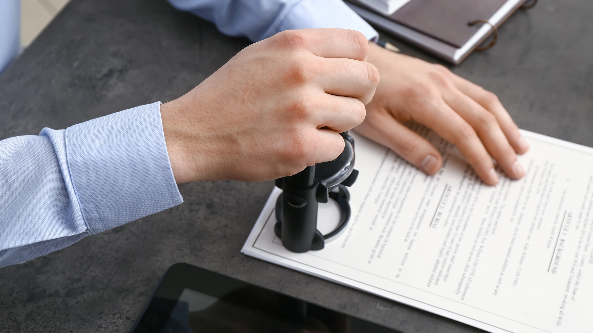 A hand pressing a black stamp onto a printed document on a dark desk while the other hand rests on the page, with a tablet and a notebook nearby