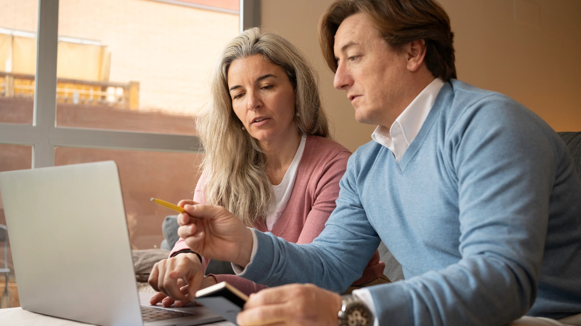 A woman and a man sit side by side looking at an open laptop, and the man holds a yellow pencil