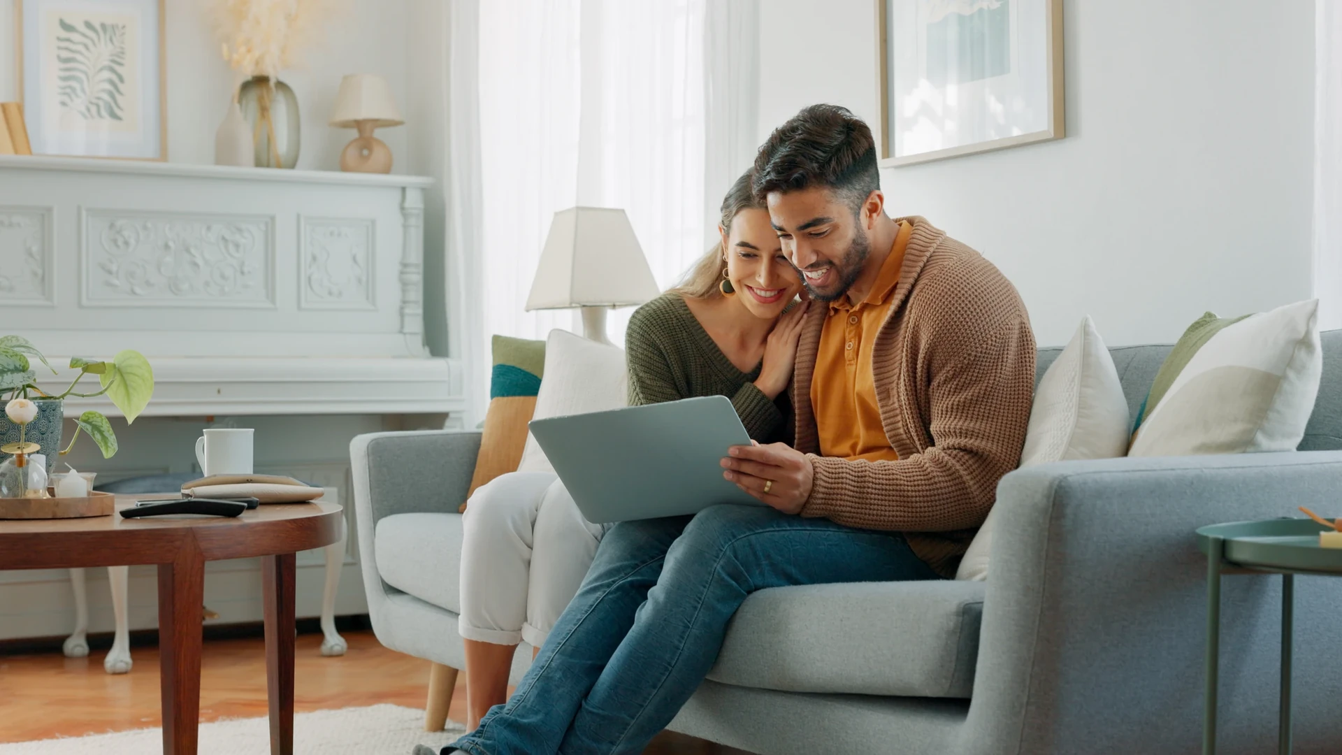 A smiling couple sit close together on a gray sofa in a bright living room, looking at a laptop on the man's lap