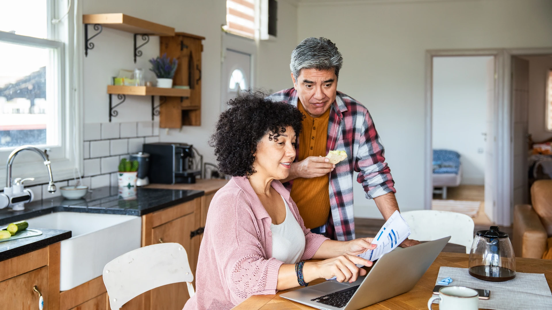 A woman holds a printed form and uses a laptop at a wooden kitchen table while a man in a plaid shirt stands behind her eating a snack