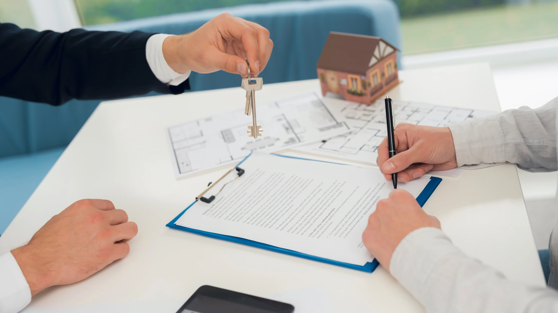 One person holds up a set of keys while another signs a document on a clipboard beside floor plans and a small model house