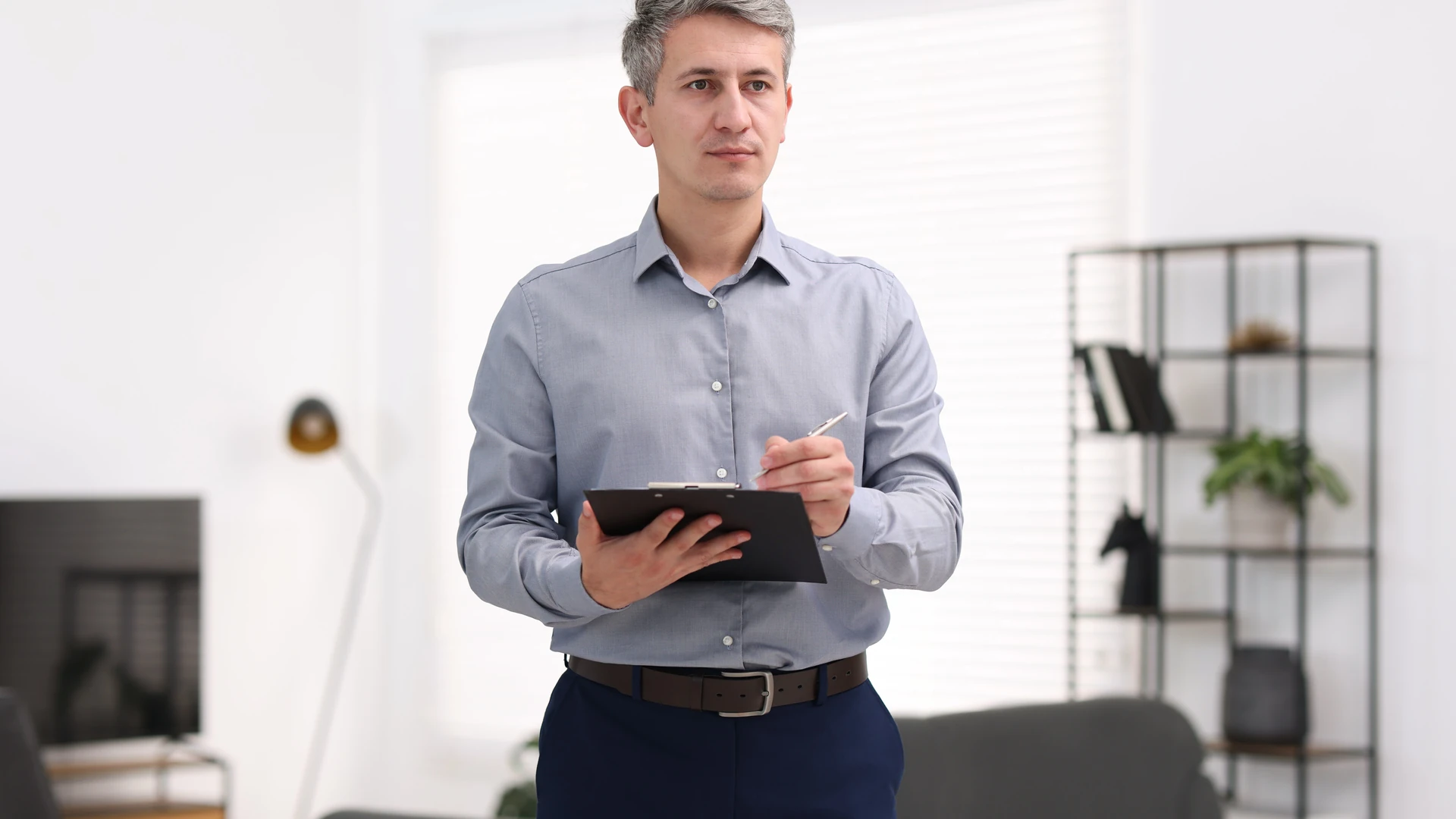 Contact Us image by Fellowship Home Loans A man in a gray button-up shirt stands in a living room holding a clipboard and a pen